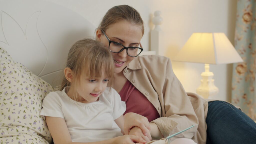 Une mère et sa fille lisent un livre au lit à la lumière d’une lampe de chevet